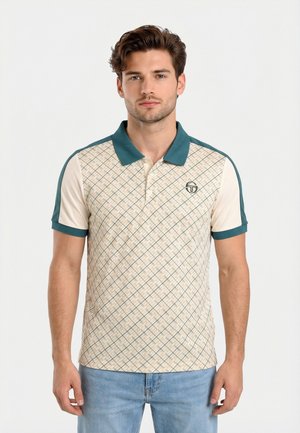 Hombre joven con cabello corto y ondulado que lleva una camisa polo beige con cuello verde y patrón geométrico, combinada con jeans azul claro, de pie frente a un fondo blanco.