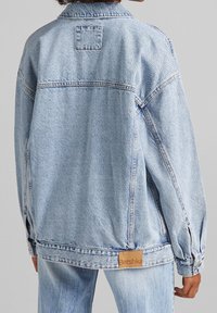 Ljusblå denimjacka med lös passform, spetsig krage och slappa ärmar. Har en liten rektangulär patch med "Bershka"-logotyp.