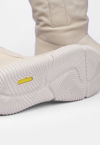 Bota de cuero beige claro con un eje ligeramente arrugado y una suela de goma blanca texturizada que presenta un patrón ondulado y un logo amarillo.
