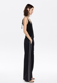 Schwarzes, funkelndes Camisole mit verstellbaren Trägern, kombiniert mit hochgeschnittenen schwarzen Hosen mit seitlichen weißen Streifen und Taschen. Flache Sandalen.