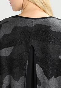 Černý a šedý pletený svetr s texturovaným maskáčovým vzorem, kulatým výstřihem a zadním rozparkem, který odhaluje černou spodní vrstvu.