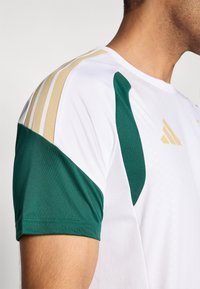 Biele športové tričko s zelenými a zlatými akcentmi, tromi zlatými pásmi na ramene, priliehavým strihom a textúrovaným materiálom.