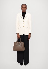 Kremfarget blazer med gullknapper, iført over en svart topp. Modellen holder en taupe håndveske med sølvfarge detaljer. Sorte vide bukser.