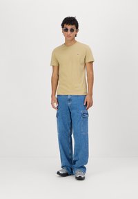 Tricou din bumbac de culoare bej, cu guler rotund, asociat cu pantaloni cargo din denim albastru, având buzunare laterale, și pantofi gri. Ochelari de soare rotunzi remarcabili purtați.