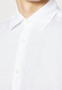 Camicia bianca di lino con colletto strutturato e bottoni visibili, realizzata in un tessuto leggermente testurizzato per un aspetto leggero.