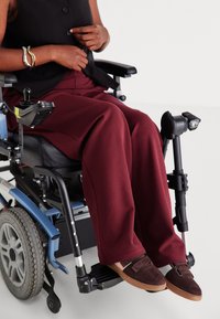 Pantalons bordeaux à jambes larges en tissu doux, assortis à des chaussures en daim marron sur un fauteuil roulant. Haut noir à boutons partiellement visible.