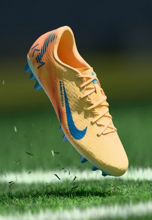 Chaussure de football Nike jaune et orange avec logo bleu et crampons soulevant de l'herbe sur un terrain en gazon synthétique vert.