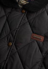 Veste noire matelassée avec un col côtelé, des boutons-pression et une étiquette de marque en cuir marron. Texture lisse avec un matelassage en motif de losanges.