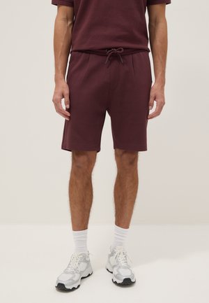 Man draagt donker bordeauxrode shorts en bijpassend T-shirt, witte sportsokken en witte sportschoenen, staand tegen een effen achtergrond.