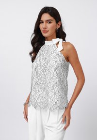 White halter top with black floral lace design, scalloped hem, and bow tie detail at neck. Worn over light-colored trousers.