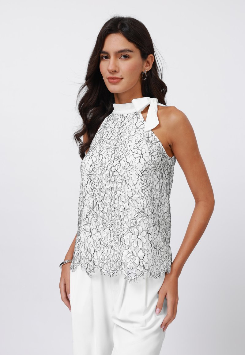 White halter top with black floral lace design, scalloped hem, and bow tie detail at neck. Worn over light-colored trousers.