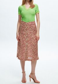 Donna che indossa un top lime verde a maniche corte con bordo in pizzo, una gonna midi rossa e beige a fantasia con bottoni, e sandali con tacco nudo.