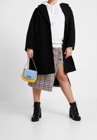 Abrigo negro, sudadera blanca, falda estampada con abertura, botas negras gruesas con cordones y un bolso bicolor con frente azul y base amarilla.