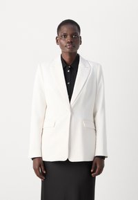 Weißer taillierter Blazer mit strukturiertem Design, Reverskragen und Fronttaschen. Über einem schwarzen Hemd mit Knöpfen getragen. Glatte Textur.