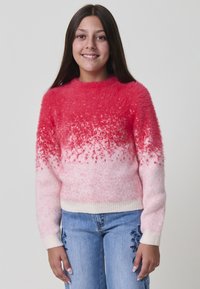 Fille souriante avec de longs cheveux bruns portant un pull en dégradé rose et rouge, ainsi que des jeans bleus avec des broderies florales, se tenant devant un fond blanc.