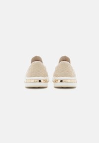 Beige slip-on-skor med texturerad överdel med geometriska mönster, guldfärgad klack och vit gummisula.