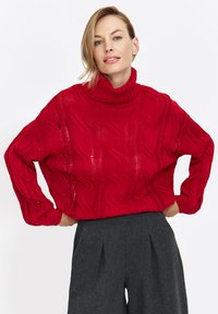 Czerwony sweter z dzianiny, z kołnierzem typu golf, z wzorem w warkocze i luźnym krojem. Połączony z szarymi spodniami o wysokim stanie.