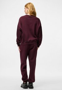 Kobieta o blond włosach ubrana w dopasowaną ciemnofioletową bluzę i spodnie dresowe, stojąca tyłem na białym tle.