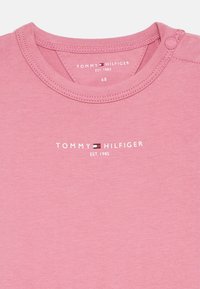 Rosa bomulls t-skjorte med rund hals, trykknappedetalj, og trykt "TOMMY HILFIGER EST. 1985" logo sentrert under halslinjen.