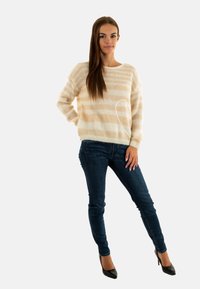 Maglione a righe beige e crema in morbido jersey, abbinato a jeans slim fit blu scuro e tacchi neri. Collo rotondo semplice e maniche lunghe.