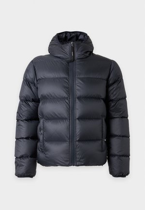 Homme portant une doudoune noire à capuche sur une chemise noire et un t-shirt blanc, associée à un pantalon gris et des chaussures marron foncé.