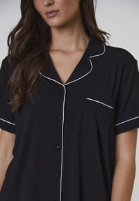 Sorte kortærmede pyjamasoberdel med hvid piping, knaplukning foran og en enkelt brystlomme. Lavet af et blødt ribbet stof.