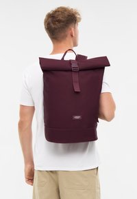 Zaino roll-top in tessuto bordeaux, con spallacci regolabili, manico superiore e una patch con il marchio vicino alla parte inferiore. Texture liscia.