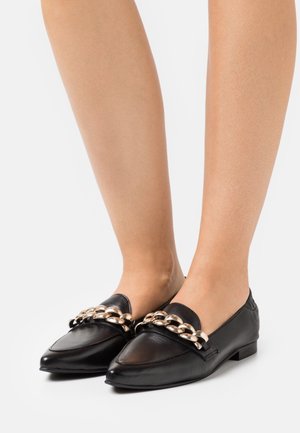 Mocassins en cuir noirs à bout pointu avec un épais détail en chaîne dorée, portés pieds nus sur un fond blanc.