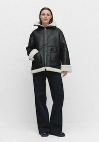 Svart läderjacka med vit fårskinnsdetalj, dragkedjestängning och två framfickor. Modellen har på sig mörka vida byxor och svarta skor.