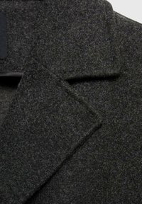 Grijze wolblendjas met een gestructureerde stof, met een aangeknipte kraag en een hoekig reversontwerp. Close-upweergave van het materiaal en de stiksels.