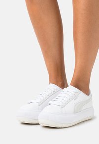 Zapatillas de plataforma blancas de Puma con cordones, llevadas sobre piernas bronceadas, contra un fondo claro y liso.