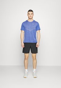 Maglietta atletica blu a maniche corte con un motivo discreto e il logo Nike, abbinata a pantaloni corti neri e scarpe da ginnastica grigio chiaro.