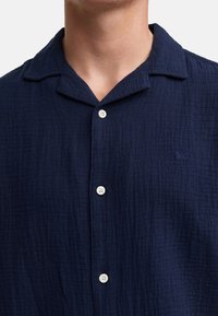Camisa azul marino de botón hecha de tejido texturizado con un cuello relajado, que presenta botones blancos y un pequeño logo azul en el lado izquierdo del pecho.