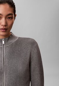 Szary prążkowany sweter z wysokim kołnierzem i zamkiem błyskawicznym, o teksturowanym wzorze tkaniny i wyraźnych szwach. Miękki dzianinowy materiał.