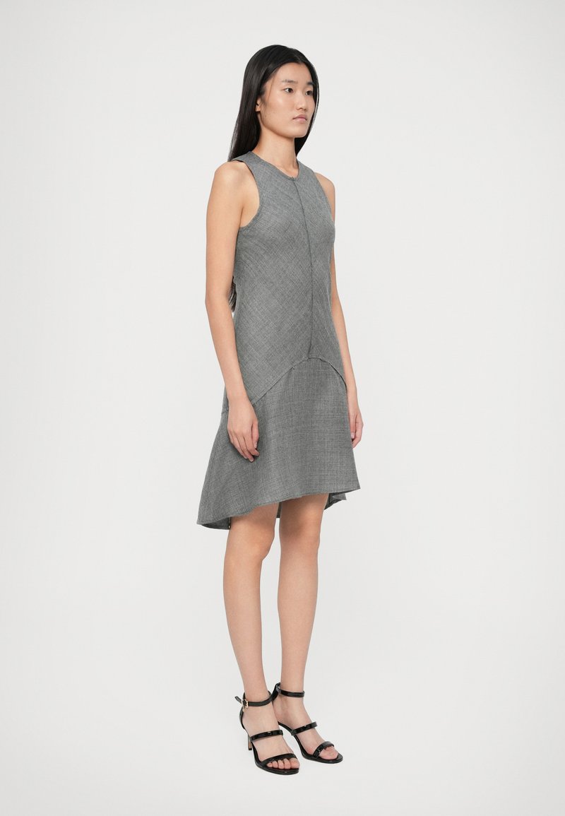 Femme debout portant une robe sans manches grise texturée avec un ourlet asymétrique et des sandales à talons hauts noires à lanières sur fond blanc.
