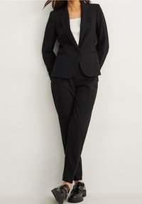 Costume noir sur mesure comprenant un blazer ajusté avec une fermeture à un bouton, un pantalon étroit et des mocassins noirs brillants. Texture de tissu lisse.