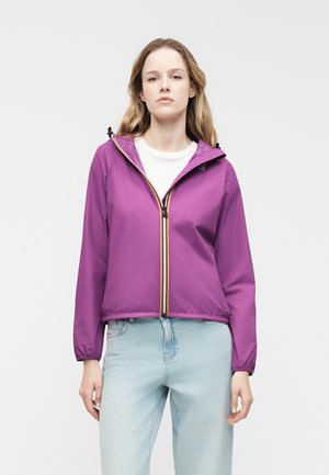 Νέα γυναίκα που φοράει μωβ ζακέτα με κουκούλα και φερμουάρ πάνω από λευκό πουκάμισο και ανοιχτόχρωμα μπλε τζιν, στέκεται μπροστά σε απλό ανοιχτό γκρι φόντο.