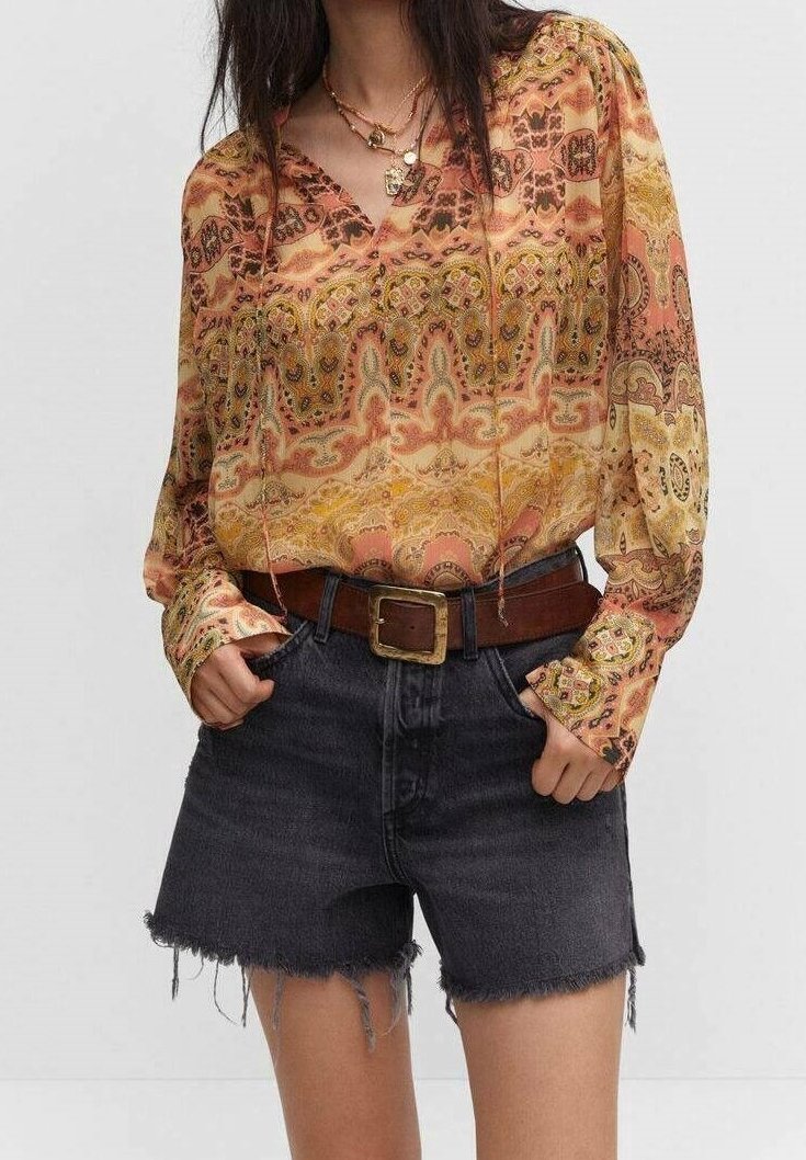 Langärmlige Bluse mit einem bunten Paisley-Muster in warmen Tönen, kombiniert mit dunklen Denim-Shorts mit ausgefransten Kanten und einem braunen Gürtel.