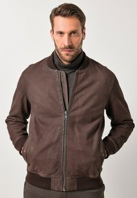 Uomo che indossa una giacca di pelle marrone con zip e un dolcevita nero, in piedi con le mani in tasca contro uno sfondo chiaro e semplice.