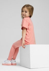 T-shirt rosa chiaro e leggings abbinati indossati da un bambino, seduto su un cubo bianco. Le scarpe sono bianche con dettagli pastello e suole a piattaforma.