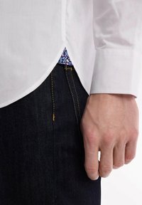 Chemise blanche à manches longues avec un ourlet arrondi, révélant un motif floral en dessous. Associée à un jean en denim foncé avec des coutures jaunes.