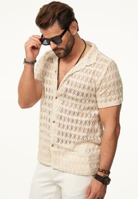 Beige, offen gestricktes Kurzarmhemd mit Kragen, verziert mit dekorativen Mustern und Knopfleiste, kombiniert mit Sonnenbrille und Armbändern.