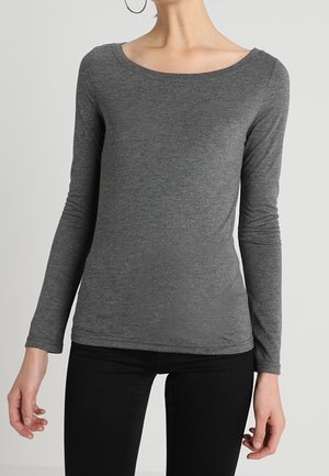 Femme portant une chemise grise cintrée à manches longues et un pantalon noir, avec de grandes boucles d'oreilles, debout devant un fond clair uni.