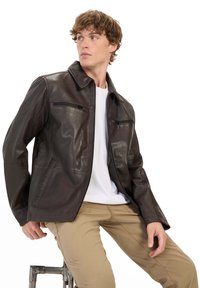 Bruine leren jack met een ritssluiting aan de voorkant, twee ritszakken op de borst en een notch-kraag, gecombineerd met een wit shirt en beige broek.