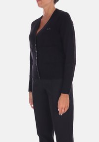 Cardigan nero realizzato in tessuto morbido, con scollo a V, chiusura con bottoni e una tasca anteriore. Presenta polsini e orlo a costine per un tocco di texture in più.
