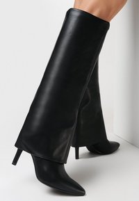 Bottes noires montantes en cuir lisse, avec un bout pointu et un talon aiguille. Le design est ajusté avec une finition lisse.