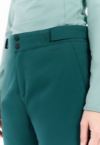 Teal outdoorbroek met een gladde textuur. Kenmerken zijn onder andere een platte tailleband met een klittenbandsluiting en twee zwarte knopen, en een subtiele merkl logo.