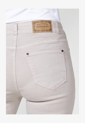 Jeans color beige claro con una textura suave, que presentan una etiqueta de marca marrón en la cintura y costuras decorativas en los bolsillos traseros.