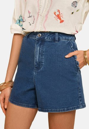 Højtaljede blå denimshorts med knap og lynlås, kombineret med en mønstret hvid bluse og guldarmbånd på håndleddet.