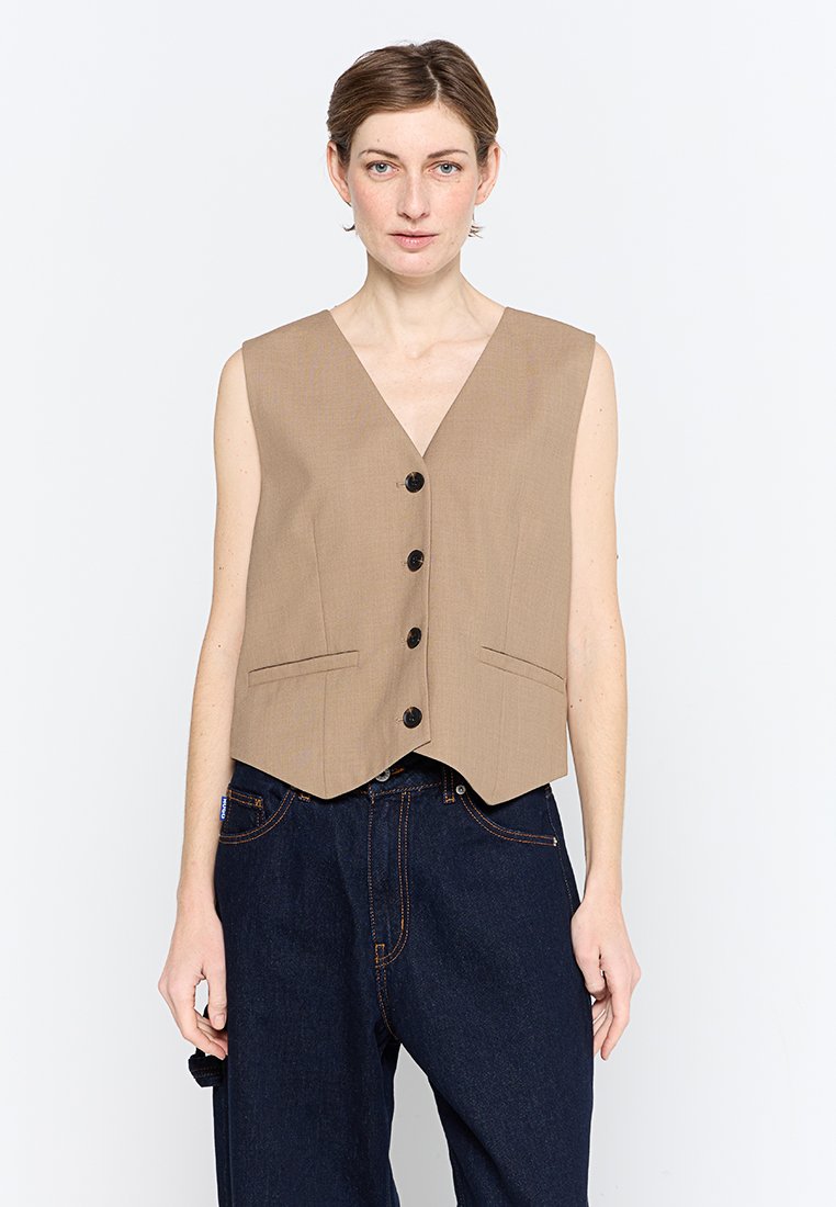 Beige mouwloos vest met een V-halsontwerp, voorzien van voorknopen en twee zakken. Gemaakt van gladde stof, getailleerde pasvorm met een cropped zoom.